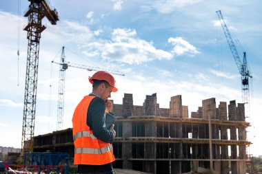 a young strong smart worker in overalls and helmet stands on a new building near the crane. Construction concept