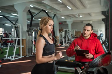 Koşu bandında koşan kişisel antrenör yardımcısı olan bir bayan sporcu. Spor salonunda insanlar spor yapıyor.
