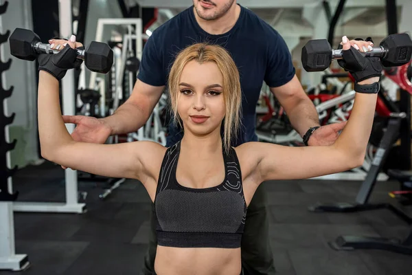 beautiful young trainer with inflated biceps trains a young cute brunette client for body health. The concept of a coach in the gym