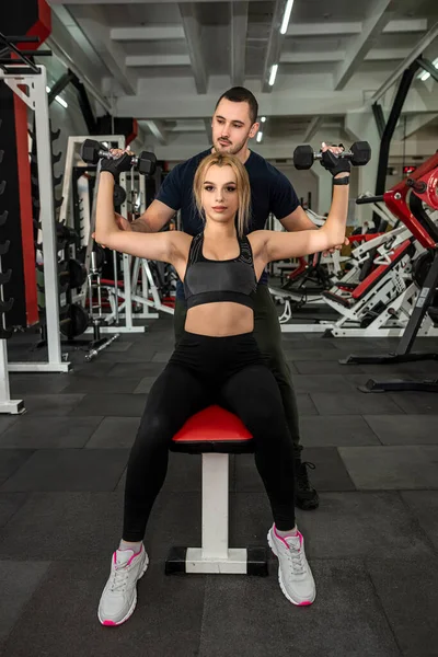 Man trainer helps her slim woman client exercising with heavy dumbbells at the gym.  Fitness and gym for health concept