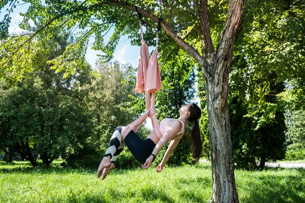 young athletically trained yoga trainer trains outdoors under a tree. The concept of yoga training on aerial canvases under a tree.