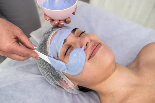 Cosmetologist applying blue mask to face woman client, rejuvenation procedure to beautiful in a beauty salon