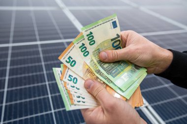 Mavi ceketli güçlü erkek elleri, Avro banknotlarını güneş panelleri önünde tutuyor. Ekonomik yeşil elektrik kavramı