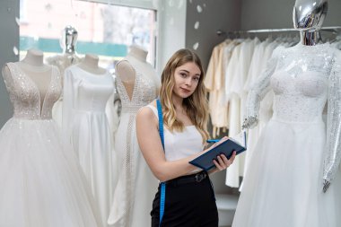 Pretty  female dressmaker with diary and pen posing near bridal wear in wedding fashion boutique