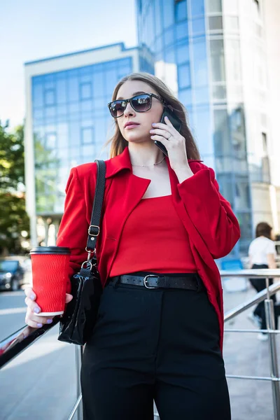 Woman in glasses and classic brand clothes holding a cup of coffee and smiling on City Center Background. Concept of woman with coffee on the street