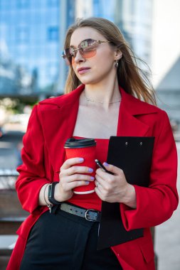 Young woman with a cup of coffee in her hands with makeup and glasses looking to the side while standing near her office. Portrait of a business girl near a glass building