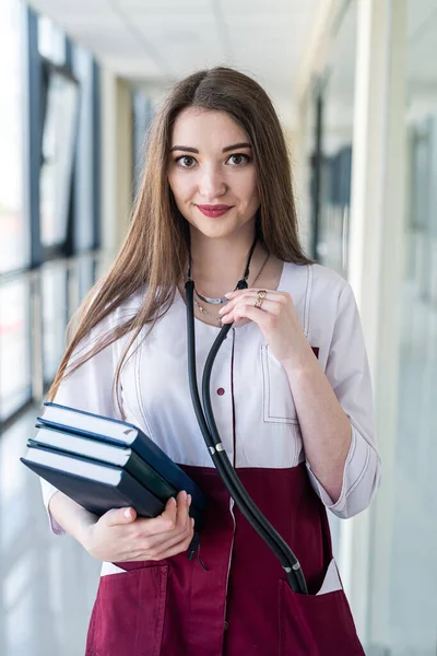 a beautiful student in a medical gown with books in her hands is standing in the corridor of the hospital. Medical student concept. Portrait of a girl