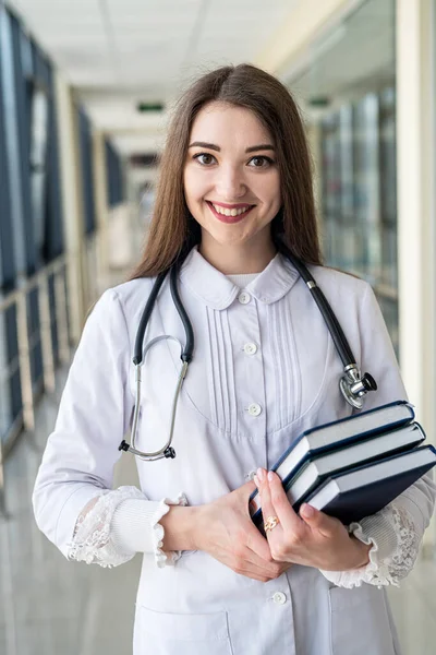 Smiling female doctor holding a book or magazine and walking down the med corridor to examine patients. Portrait of a female doctor. Medicine saves lives. A beautiful nurse