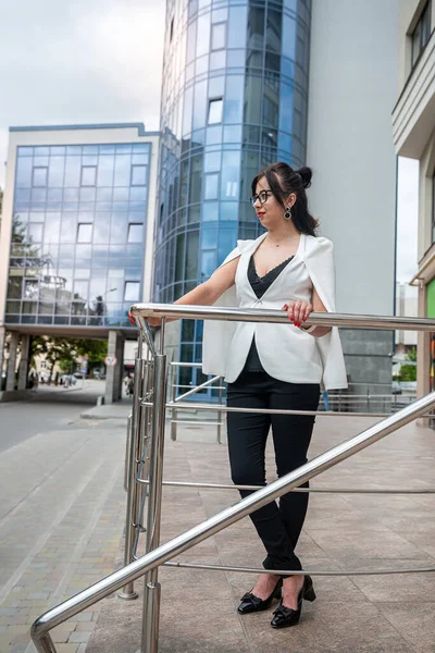 a businesswoman in a business style suit stands with a bag in her hands against the background of a modern office center. The concept of a business woman in the middle of the city