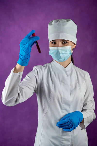 Isolated female doctor in medical mask and blue gloves holding test tube with blood sample for coronavirus test on plain background. Respiratory disease. Concept of medicine