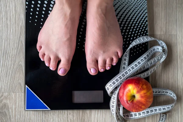 Cropped image of woman standing on scales against measuring tape and apple. We wrap the legs with a tape measure. body weight measurements. female feet