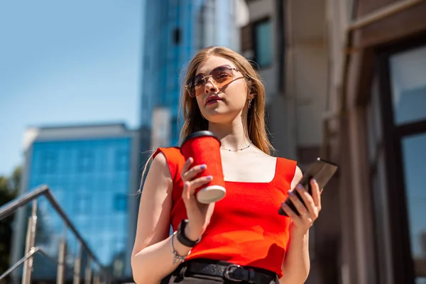 A business woman with a cup of coffee is talking on her smartphone while standing in the middle of the city center. The concept of a woman who earns money