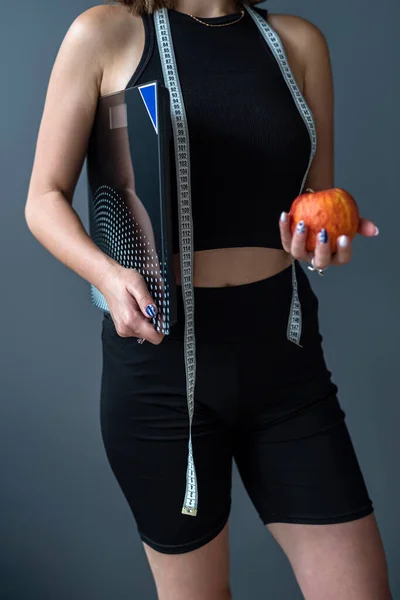 Young happy sport sports fitness trainer instructor woman wearing black sports suit hold apple measuring tape weight isolated on gray background. Sport workout concept