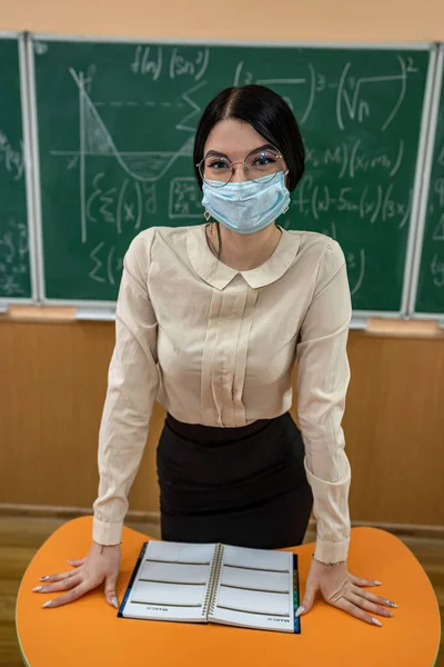 Portrait of teacher in medical mask during coronavirus epidemic standing against blackboard. Educative mathematics lesson