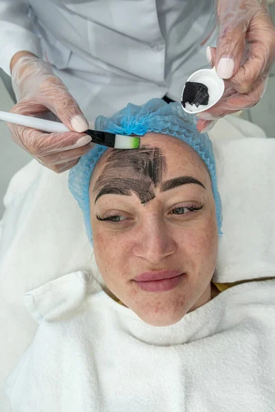 satisfied young beautiful woman uses the services of a professional cosmetologist in a beauty salon or spa. cosmetologist applied a white mask with a brush. Top view. cosmetology