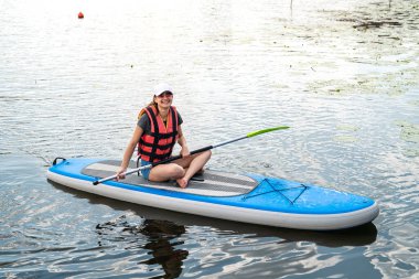 smiling woman sitting on sup board with paddle. resting and sunbathing on a nature