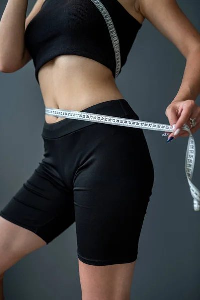 waist view of a sporty woman measuring her size with a tape. sport lifestyle concept. women\'s sizes female waist on isolated gray background. studio shooting