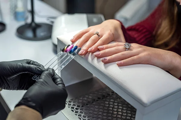 A master in rubber gloves and a young female client in a protective mask choose a color for a manicure in the interior of a beauty studio. Manicure and pedicure salon covid-19 and social distance.