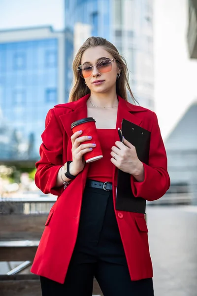 Woman in glasses and classic brand clothes holding a cup of coffee and smiling on City Center Background. Concept of woman with coffee on the street