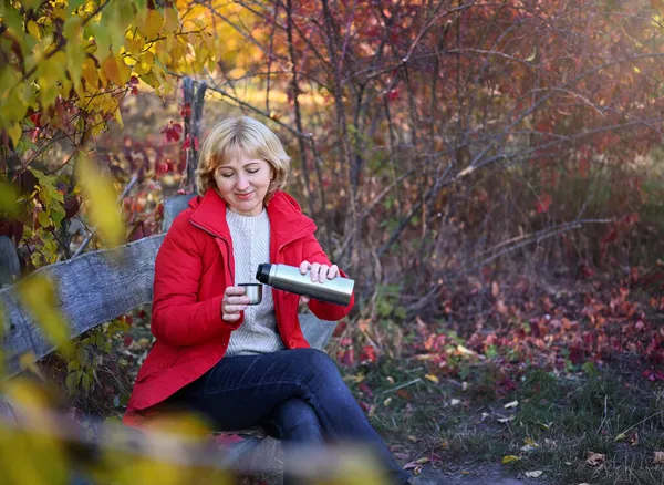 50-60 yaşlarında hoş bir kadın sonbahar parkında, ahşap bir bankta oturuyor, ısınmak için termostan çay döküyor. Kırmızı bir ceket giyiyor, bej bir kazak. Sonbahar doğası..