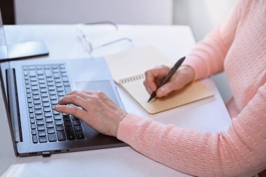 Olgun kadın evde dizüstü bilgisayar kullanıyor, deftere yazıyor. Masanın üzerinde gözlük ve cep telefonu var. Evden çalışma, çevrimiçi öğrenme ve serbest öğrenim kavramı.