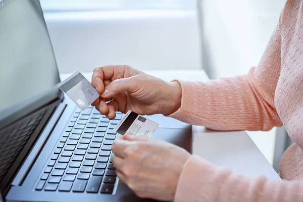 Mature woman works at home on a laptop. She is holding two credit cards in her hand. Online shopping, work from home and freelance concept. Shallow depth of field, soft focus.