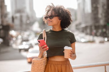 Koyu tenli, güneş gözlüklü, akıllı telefonlu genç bir kadın.