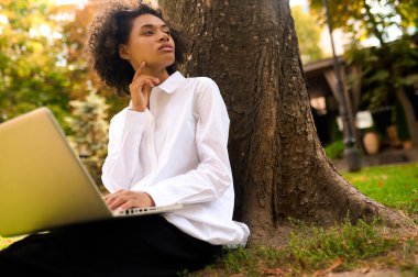Dizüstü bilgisayarı olan genç bir kadın elinde dizüstü bilgisayarla ağacın altında oturuyor.