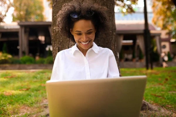 Tmavovlasá dívka sedící pod stromem s laptopem — Stock fotografie