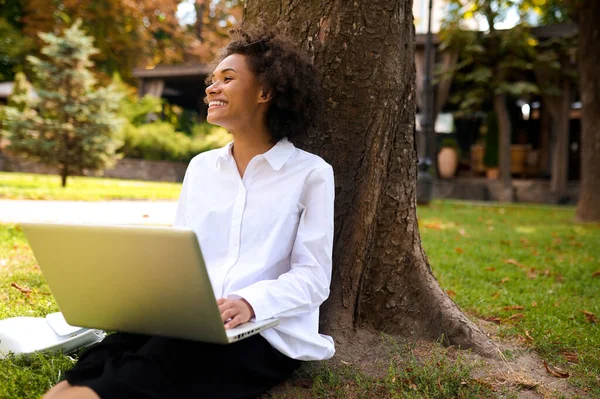 Σκούρα μαλλιά κορίτσι κάθεται κάτω από το δέντρο με ένα φορητό υπολογιστή — Φωτογραφία Αρχείου
