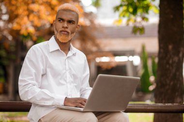 Saçları boyalı genç bir adam parkta bankta oturuyor ve dizüstü bilgisayarla çalışıyor.