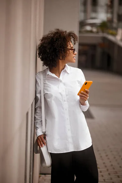 Una joven mulata mirando a un lado mientras está de pie cerca de la pared con el teléfono en las manos — Foto de Stock