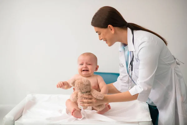 Pediatra carinhoso dando um ursinho de pelúcia para um bebê chateado — Fotografia de Stock