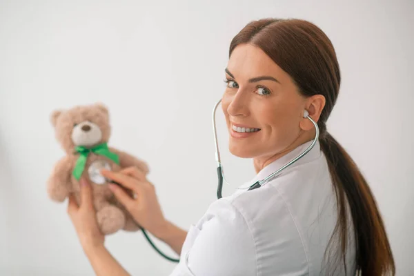 Médico sorridente realizando um procedimento de ausculta em um ursinho de pelúcia — Fotografia de Stock