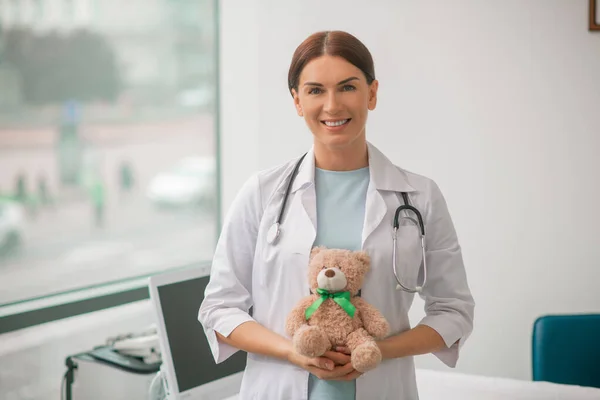 Vista frontal de uma médica alegre com um brinquedo macio e um estetoscópio em pé em seu escritório — Fotografia de Stock