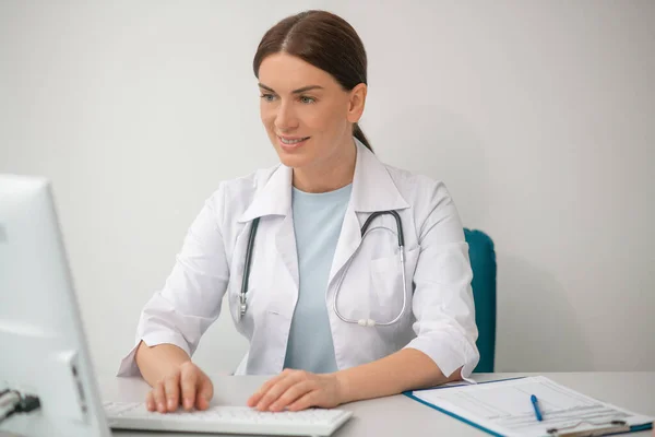 Doctora morena vestida con una bata blanca sentada en el consultorio médico — Foto de Stock