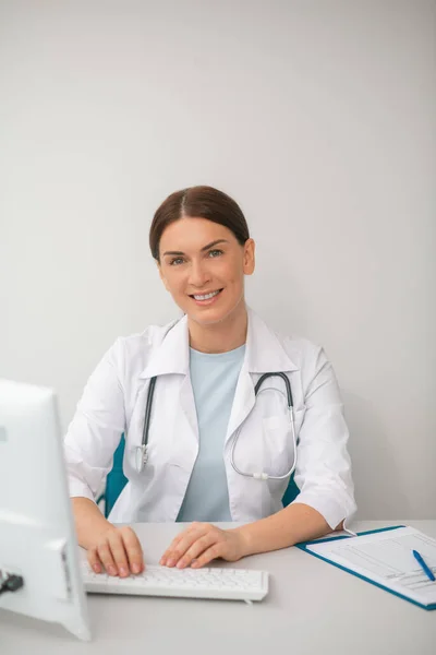Médica de cabelos escuros em um manto branco sentado no consultório médico — Fotografia de Stock