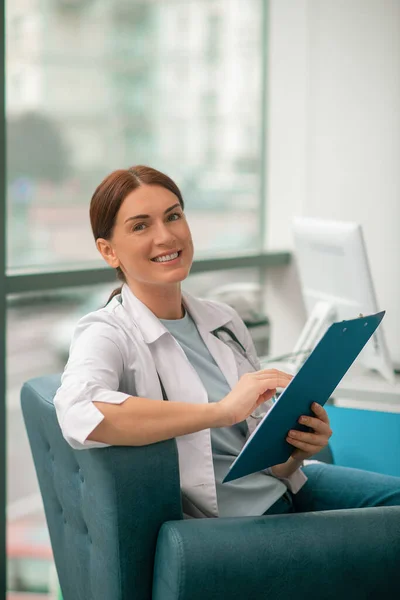 Uma mulher sorrindo em um manto branco segurando uma lista de prescrições e olhando satisfeito — Fotografia de Stock