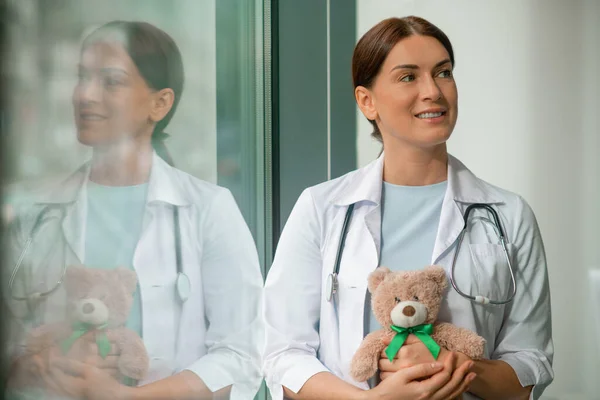 Un lindo pediatra de pie cerca de la ventana y sosteniendo un oso de peluche —  Fotos de Stock