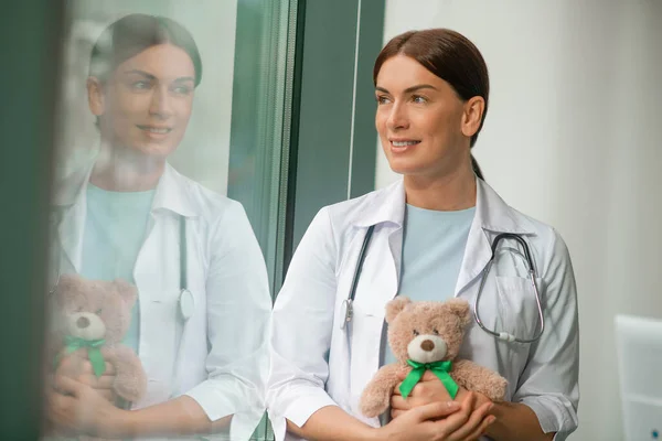 Ένα χαριτωμένο παιδίατρος στέκεται κοντά στο παράθυρο και κρατώντας ένα αρκουδάκι — Φωτογραφία Αρχείου