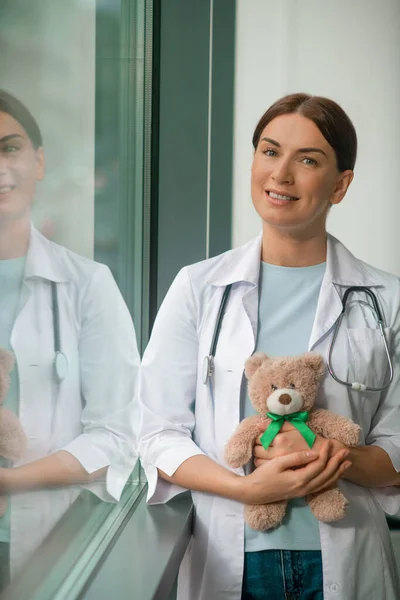Un lindo pediatra de pie cerca de la ventana y sosteniendo un oso de peluche — Foto de Stock