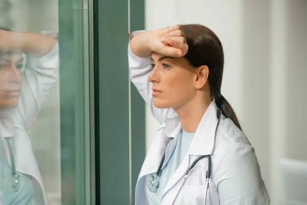 Un médico de cabello oscuro de pie cerca de la ventana y mirando pensativo — Foto de Stock