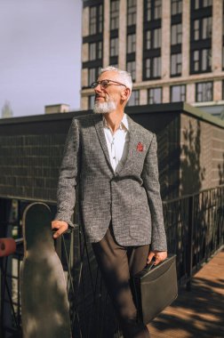 Şık, gri saçlı, olgun bir adam şehir arka planında duruyor.