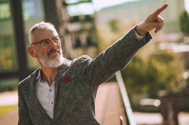Gözlüklü gri saçlı adam el hareketi yaparak yön gösteriyor.