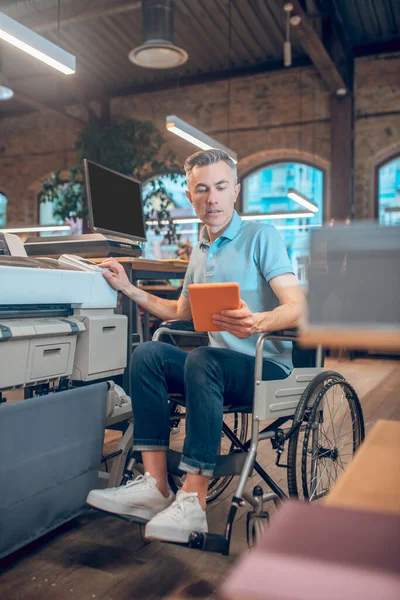 Άνδρας σε αναπηρική καρέκλα με tablet κοντά σε φωτοτυπικό μηχάνημα — Φωτογραφία Αρχείου