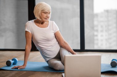 Gray-haired senior woman sitting on the floor and having an online yoga class