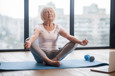 Gray-haired senior woman sitting on the floor and having an online yoga class