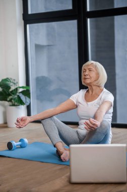 Gray-haired senior woman sitting on the floor and having an online yoga class