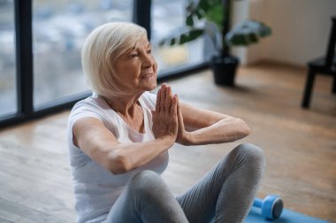 Gri saçlı yaşlı kadın yerde oturmuş meditasyon yapıyor.