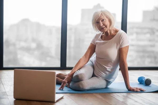 Grauhaarige Seniorin sitzt auf dem Boden und nimmt an einem Online-Yoga-Kurs teil — Stockfoto